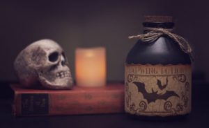 Potion in the foreground. Skull and lit candle sitting on a book in the background. 