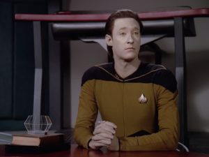 Brent Spiner as Data in Star Trek: The Next Generation. He is sitting at table with his fingers laced together. 