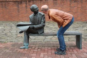 man peering at statue of other man who is reading a newspaper.