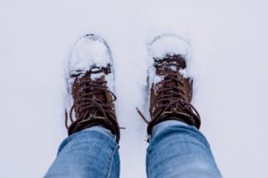 Person wearing brown boots and blue denim. The're standing on snow and their boots are caked in snow. 