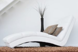 A white chaise lounge. It has two pillows on it and is sitting next to a vase filled with dead branches. 