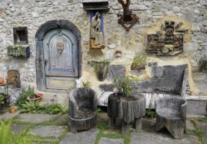 Two chairs and a table carved out of large logs of wood. They're sitting in a small garden next to a stone house that is covered with wooden carvings of people and animals. 