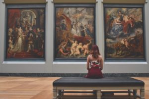 Woman sitting on a bench in an art museum looking at large paintings. 