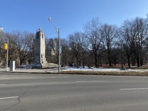 Landscape photo of a park. Image on left is of a World War I statue. There are numerous trees in the background and a road in the foreground. 