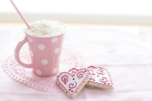 a hot chocolate topped with whipped cream sitting in a pink, polka dotted mug. There are two heart-shaped cookies sitting on a doily next to the mug. 