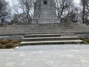 A photo of a statue in a park. There are steps leading up to the statue and the bushes around it are still dormant from the winter. 