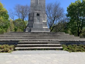 Close-up shot of a moment. There are green trees in the background and green bushes in the foreground next to the steps on the monument. 