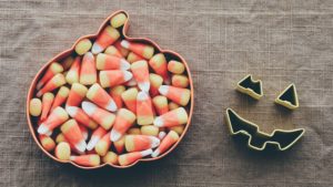 A pumpkin-shaped tin filled with candy corn. There are other pieces of tin sitting on the table next to it that look like the carved-out eyes and mouth of a pumpkin. 