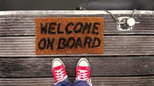 A snapshot of the legs and feet of someone wearing jeans and red sneakers. They're standing next to a "welcome on board" mat on what appears to be a wooden pier. 