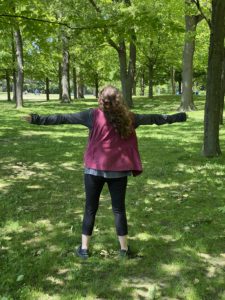 Image of the backside of a woman who is stretching out her arms and lifting her face towards the sun in a forest. 