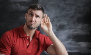 Man in red polo shirt sitting in front of chalkboard and holding his hand up to his ear as if to eavesdrop. 