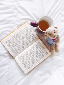 Opened book next to cup of tea and stuffed toy bear. All three items are sitting on a white bedspread. 