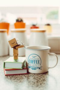 A cardboard robot standing on two stacked books while stirring a cup of coffee with a teaspoon 