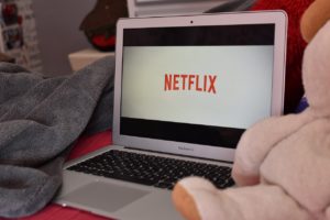 A laptop with the Netflix logo on it. The laptop is sitting on a bed and flanked by a stuffed animal and a blanket. 