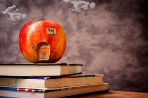 A toy apple sitting on three textbooks in front of a blackboard. The toy apple has a door, window, and lantern painted onto it so that it looks ilke a litle house. 