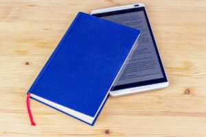 An ereader tucked inside of a hardback novel. The novel is sitting on a wooden surface, possibly a table or floor. 