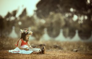 Child reading a book while sitting on brown grass and wearing a wreath of flowers in her hair outside 
