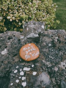 A rock painted orange that says "stay safe be kind." It is lying on a much larger, lichen-covered rock. 