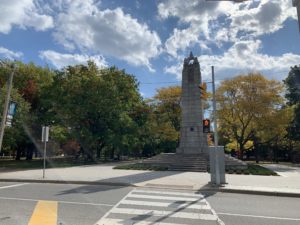 Landscape photo of a World War I monument behind a crosswalk and in front of several trees that are changing from green to yellow as autumn deepens. 