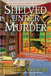 Shelved Under Murder (Blue Ridge Library Mysteries, #2)  by Victoria Gilbert book cover. Image on cover is taken from library looking out it's big picture window. There is a tree with a crime scene do not cross tape around it and many red, yellow, and orange leaves on the ground. 