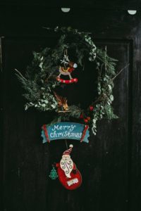 Christmas wreath with a Santa placard saying "Merry Christmas" hung from it. The wreath is hung on a slightly ominious black door.