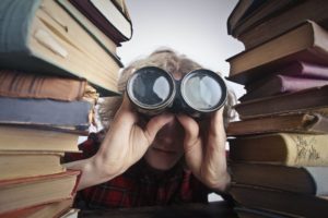 Man using binoculars while sitting between two stacks of thick, dusty books at a large wooden table