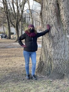 A petite woman bundled up for winter and standing next to a large tree