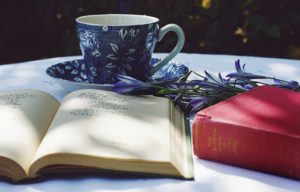 book opened on top of white table beside closed red book and run blue foliage ceramic cup on top of saucer 