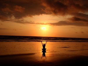 silhoutte of person sitting by a beach and raising their hands to the sky as the sun sets