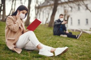 two people wearing masks, social distancing, and reading books outside at a park.
