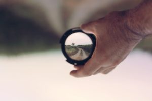 Person holding a camera lense that is focused on a dirt road through a field. A small patch of woods is in the distance just around the bend of the road. 