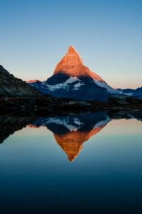 A mountain half bathed in evening sunlight. It is reflected in a perfectly-still lake at the foot of the mountain. 