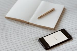 Smartphone near an empty notebook and pen. All items are sitting on a white and grey striped blanket. 