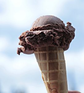 a chocolate ice cream in a cone held up against a blue and white sky 