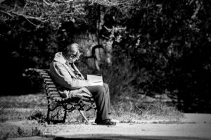 black and white photo of slumped over man reading a book while sitting on a park bench outdoors 