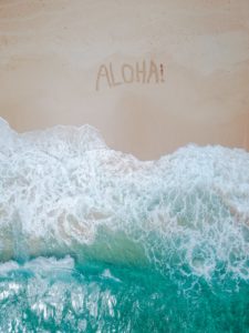 Arial shot of waves gently lapping against a large sandy beach. Someone has scratched the world aloha into the sand. 