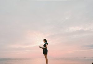 Woman standing on a beach at sunset reading a book. There is a beautiful pink sky behind her. 