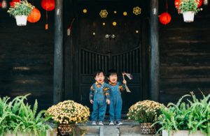 Toddler twins standing in front of a large wooden door adorned with red lanterns.