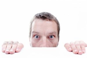 Man peeking out from behind a white desk in a white room. He looks nervous. 