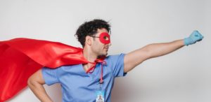 Man wearing a red superhero cape and a red eye mask holding his fist forward. He’s wearing scrubs and gloves and so is probably a healthcare worker. 
