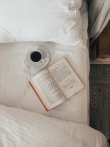 A cup of coffee and an opened book lying on a bed whose top sheet has been folded back 
