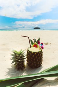 A frothy pineapple drink in a pineapple. The pineapple is sitting on sand at a tropical beach. 