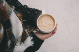 Person wearing a heavy winter coat and scarf carrying a mug of hot chocolate. 