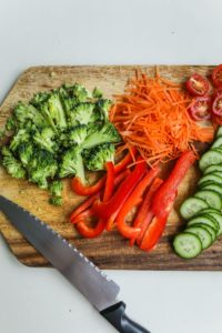 A wooden cutting board that has a knife and chopped broccoli, carrots, red pepper, cucumber, and tomatoes on it. 
