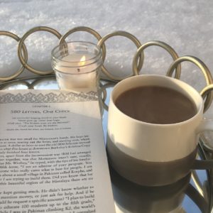 A cup of coffee, a tealight candle, and an opened book on a mirrored platter that’s lying in the snow. 