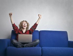 Person with curly hair and glasses cheering on a blue couch as they hold a laptop in their lap.
