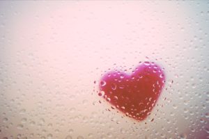 A red heart that’s been affixed to the other side of a windowpane that’s foggy and covered with rain. 
