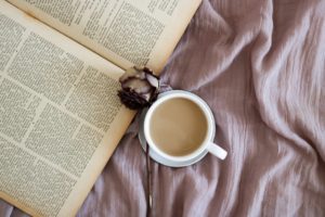 A cup of coffee, a dried rose, and an opened book lying on a light purple blanket. 