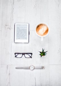 An ereader, a cup of coffee, a pair of black glasses, and a watch sitting on a wooden table. 