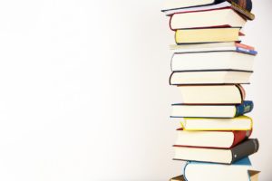 A stack of about a dozen books sitting next to a white wall. All of the book’s spines are facing away from the viewer so we don’t know their topics or authors. 